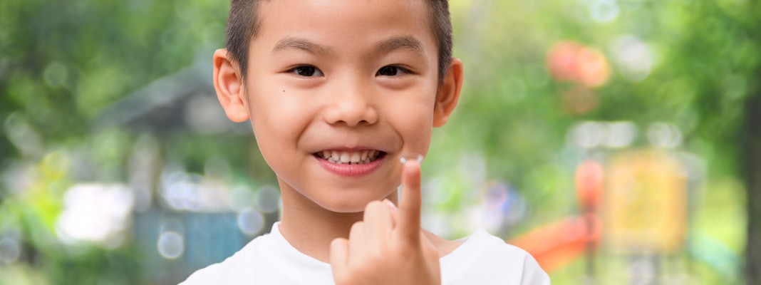 Child holding a contact lens on their fingertip while smiling outdoors.