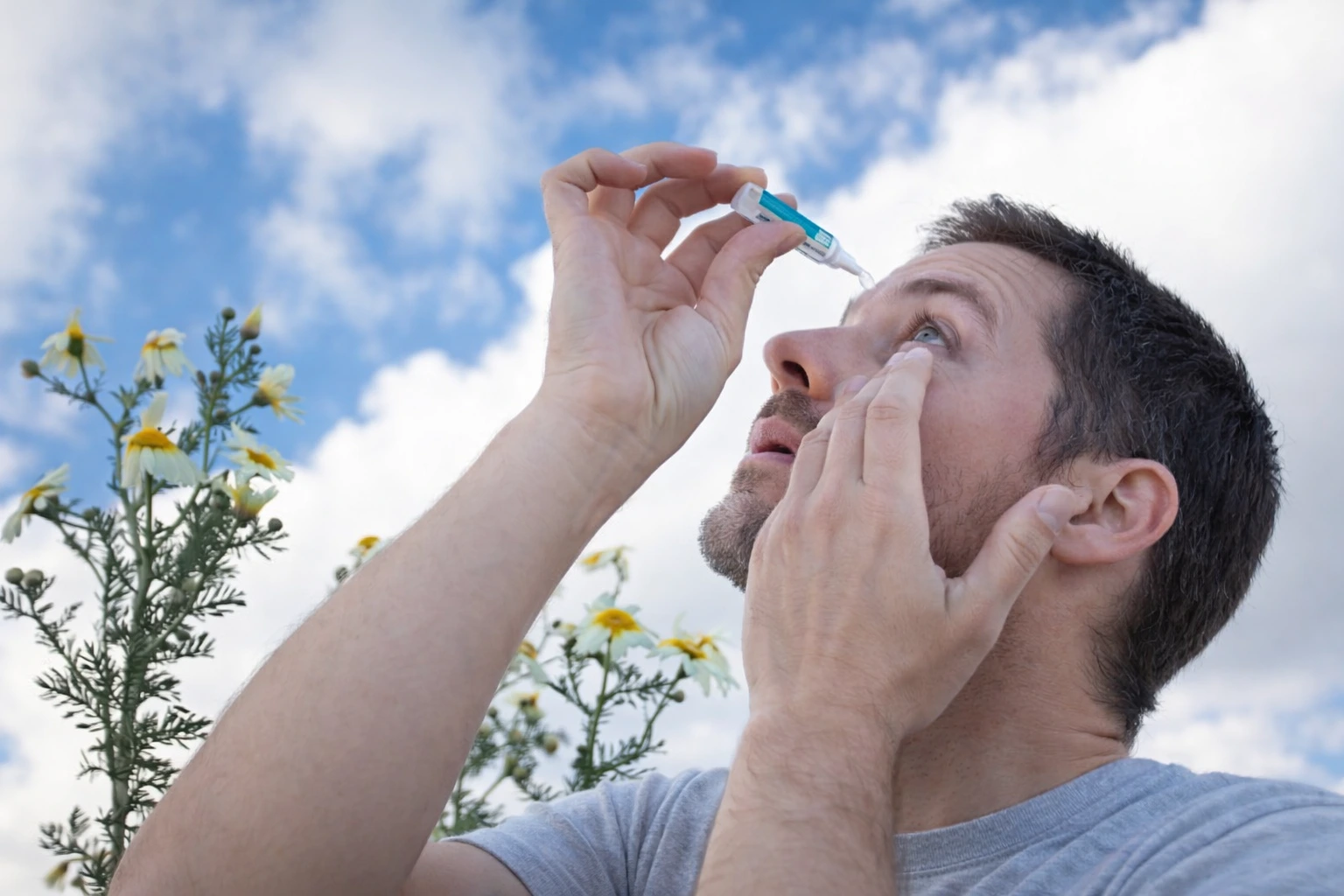 A man outdoors in spring applies lubricating eye drops under a blue sky, a common relief step for contact lens for spring allergies irritation and dryness.