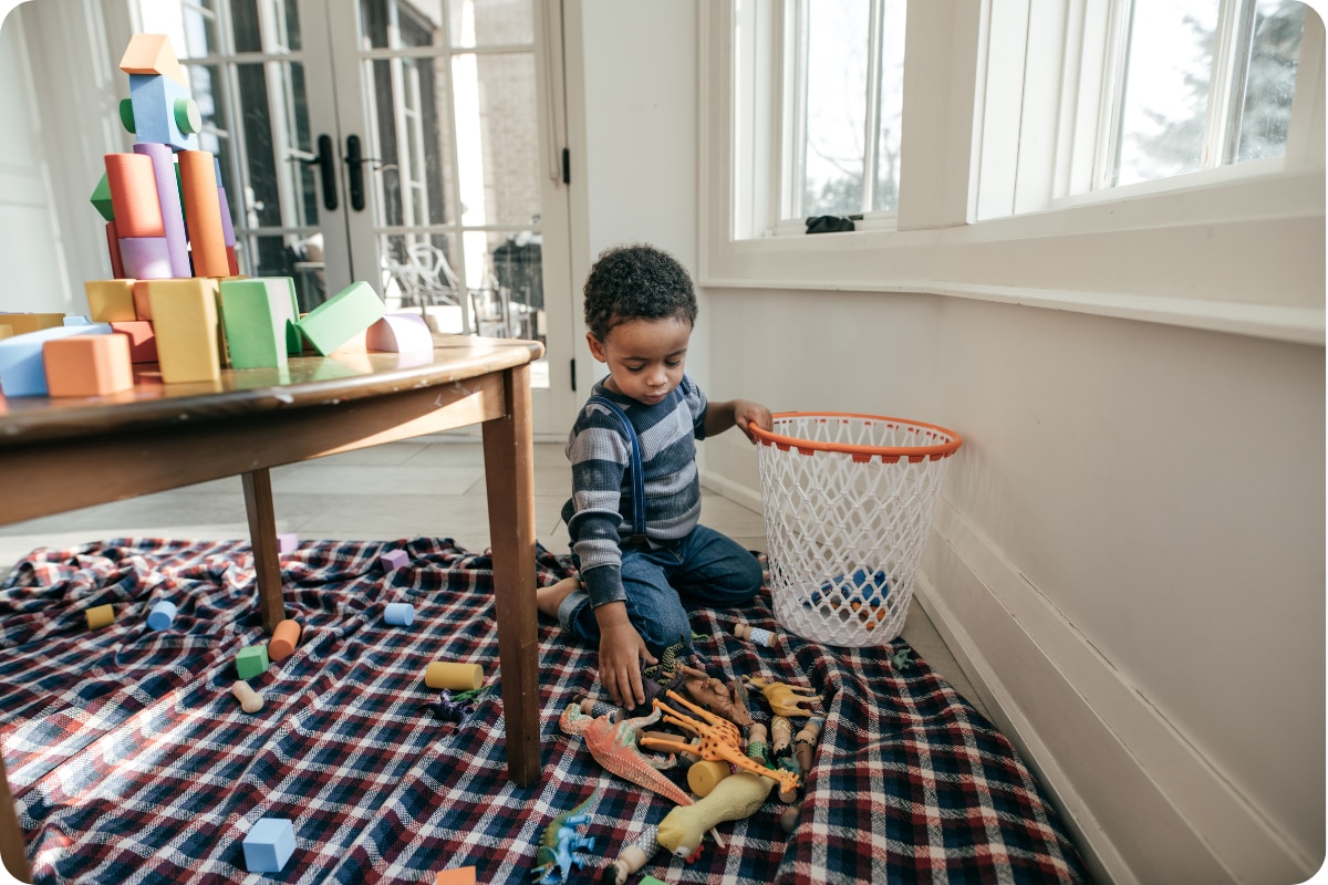 Child sitting on a blanket indoors, sorting toy animals and blocks into a small basket near a table.