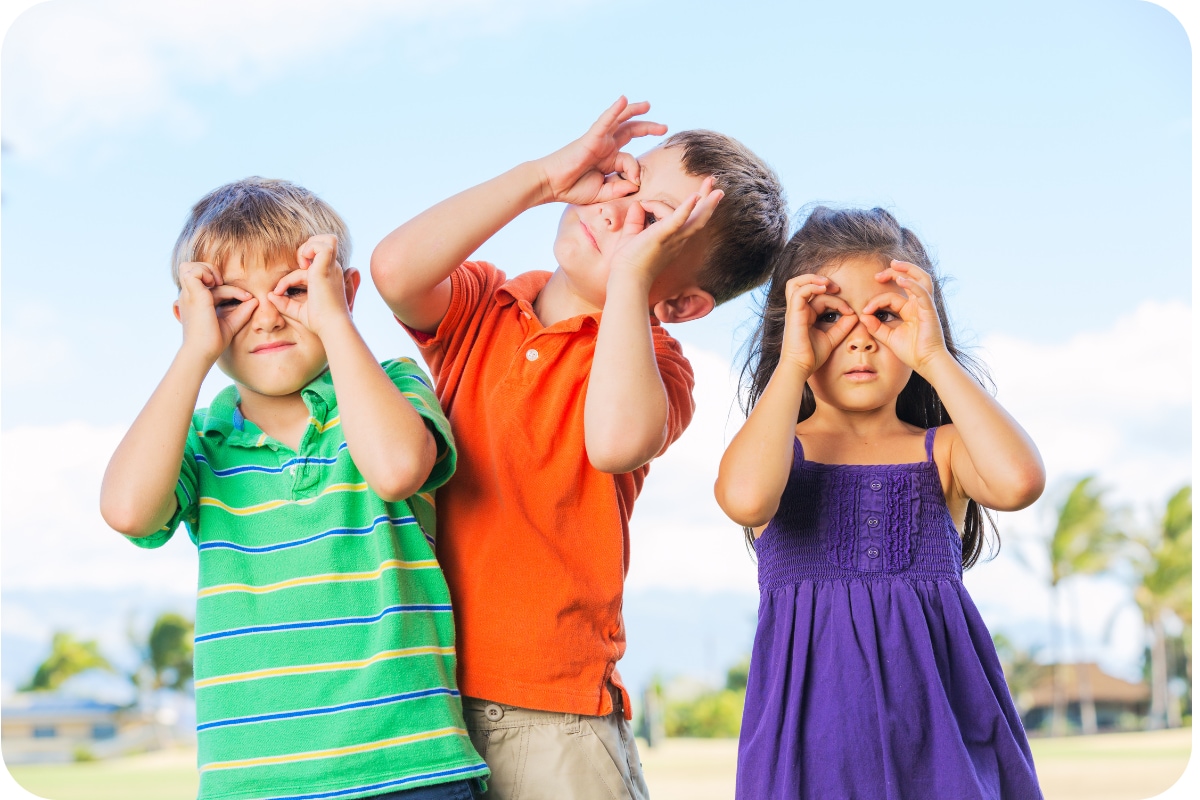 Three young children outdoors making binocular shapes with their hands over their eyes while looking ahead.