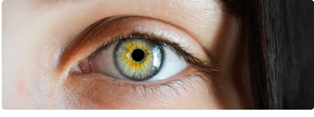 Close-up of a human eye with a green-hazel iris, visible eyelashes, and natural skin texture in soft light.