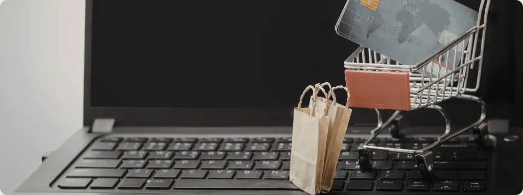 Mini shopping cart with a credit card and small paper bags sitting on a laptop keyboard, representing online shopping.