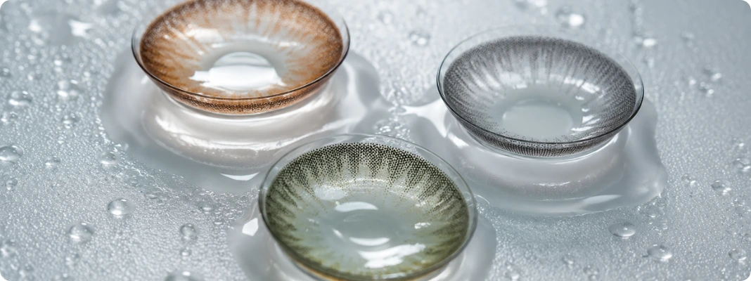 Three tinted contact lenses in honey brown, gray, and green arranged on frosted glass with water droplets in a clean macro shot.