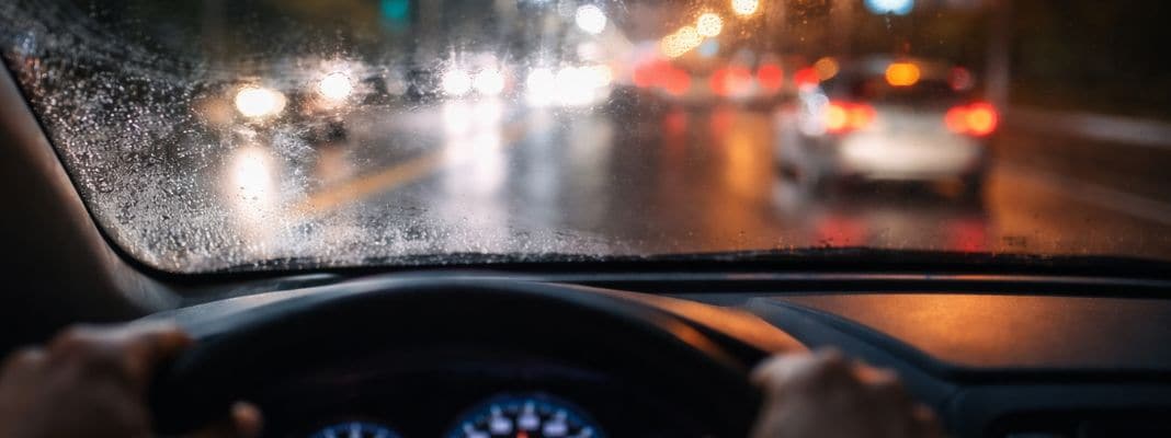 Driver’s view through a rain-speckled windshield with blurred headlights and streetlights at night.