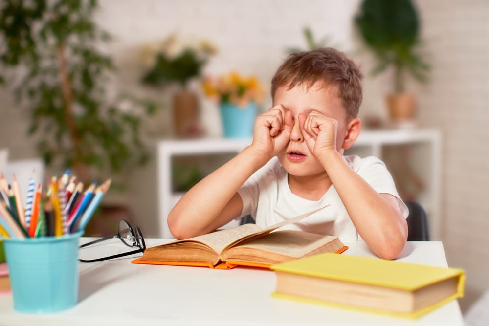 Warning Signs Your Child May Have a Vision Problem: child rubbing both eyes while reading at a desk with books and school supplies nearby.
